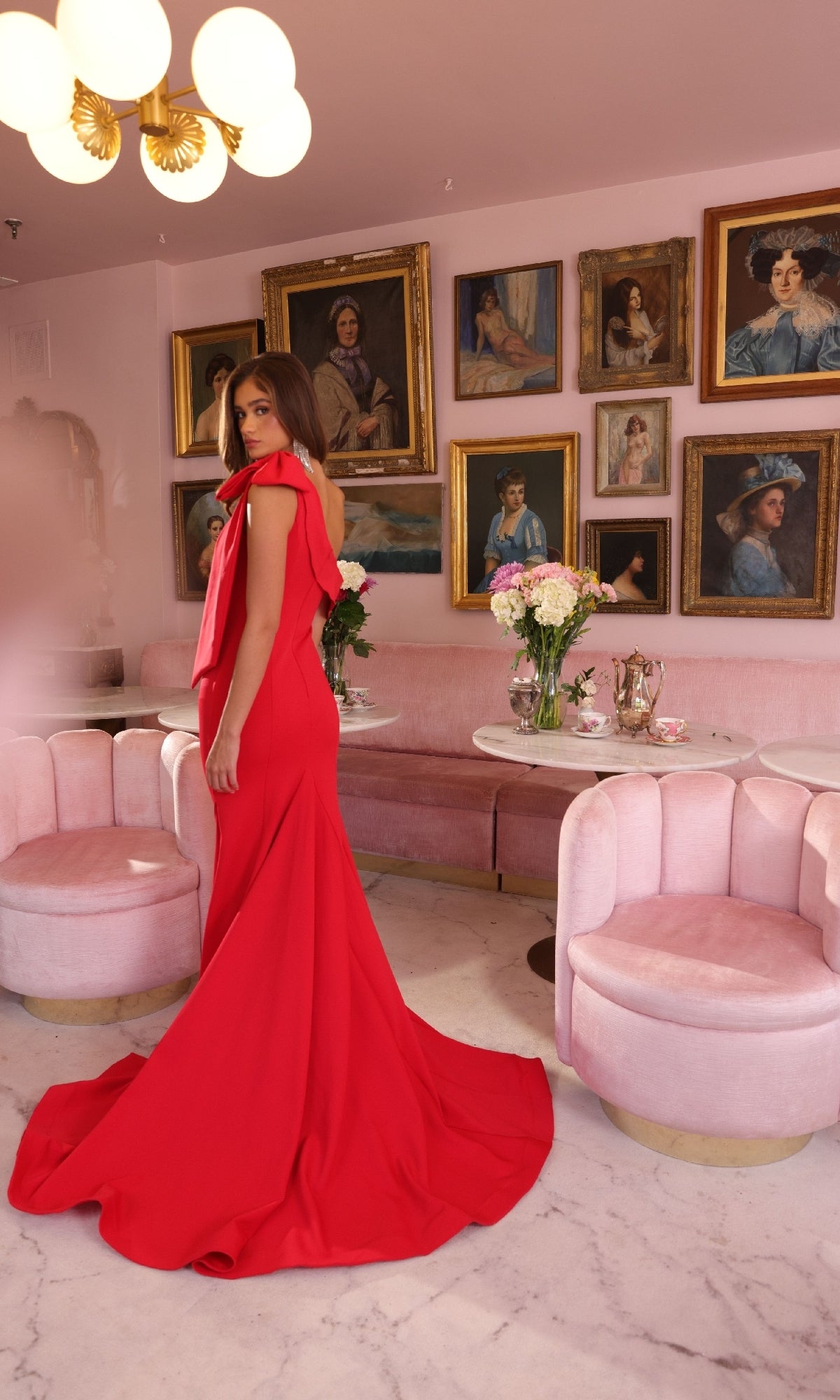 A woman wears the Ava Presley 40023 one-shoulder long formal dress in an elegant room with pink velvet furniture, marble floors, floral arrangements, and portrait paintings on the wall.